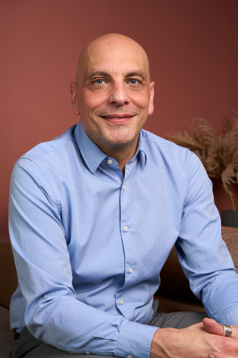 Portrait corporate d'un collaborateur chauve et souriant en chemise bleue sur fond terracotta, cabinet de conseil Paris
